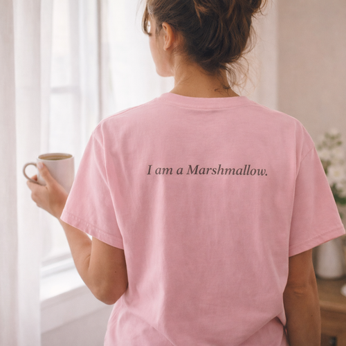 Person wearing a pink t-shirt with 'I am a Marshmallow' text, holding a mug indoors.