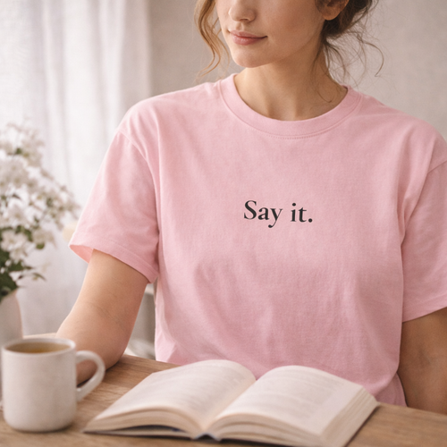 Person wearing a pink t-shirt with 'Say it.' text, sitting at a table with a book and mug.